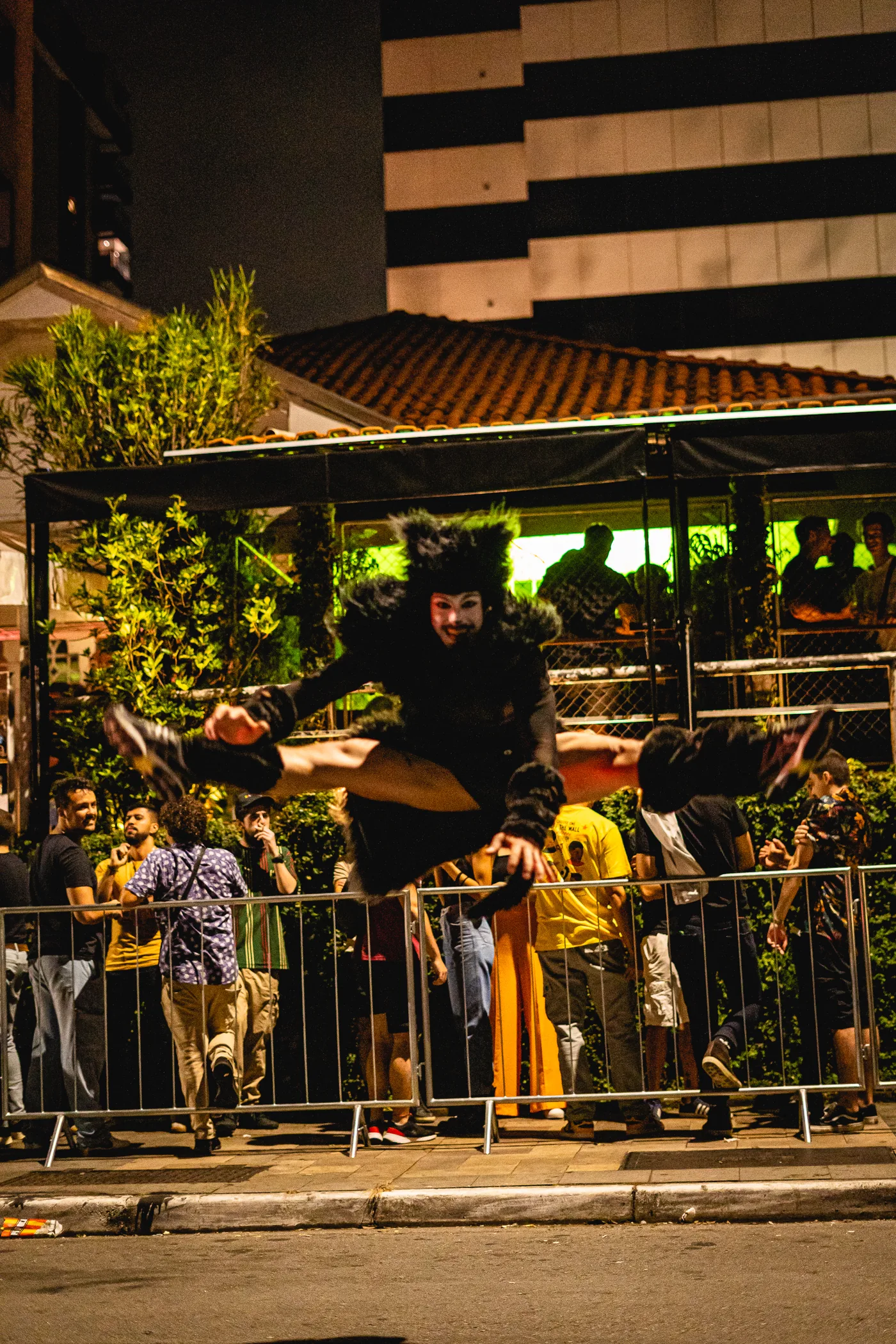O gato preto do musical em pleno salto na rua, diante da grade com a fila