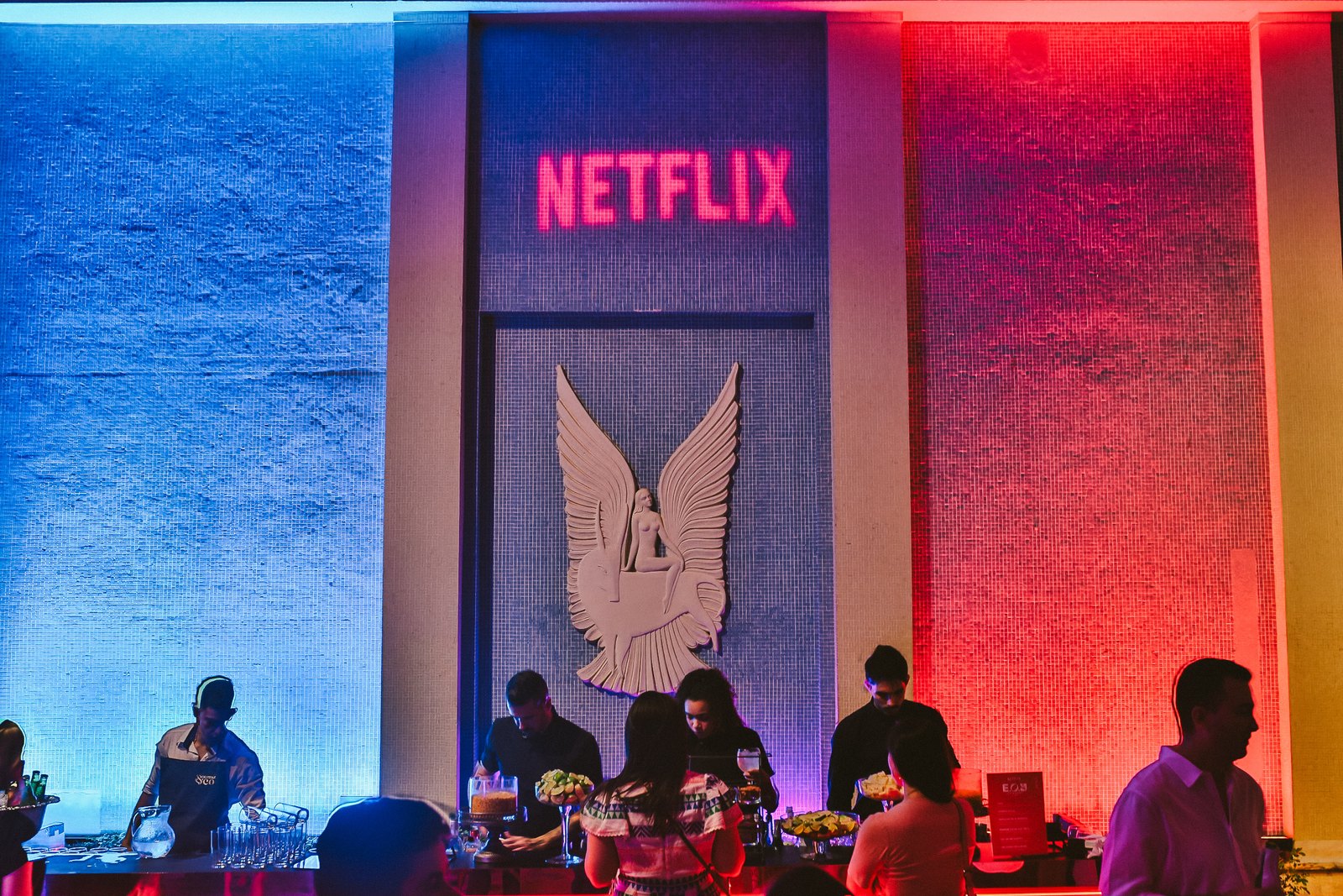 Bar principal cheio, sob o letreiro Netflix e a luz vermelha e azul